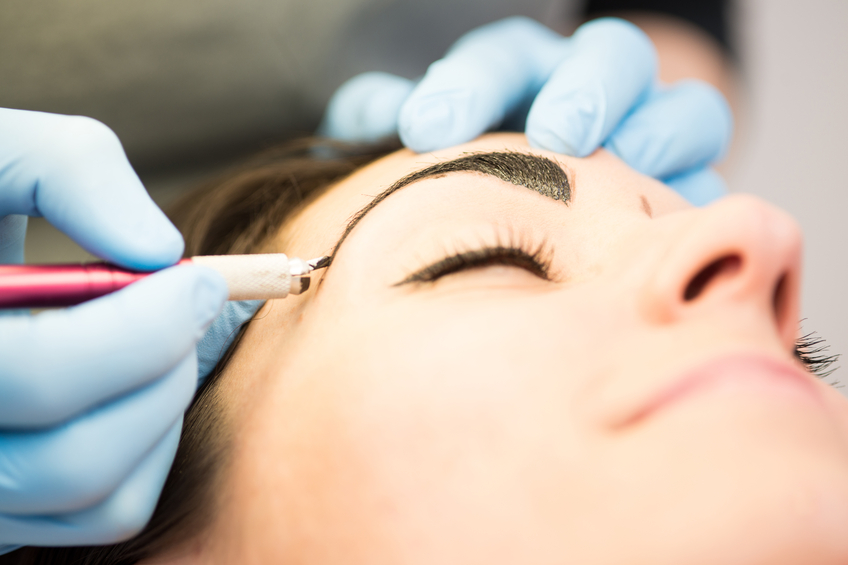 Close-up of a beautiful satisfied young woman with perfect clean fresh skin and hazelnut eyes, having her eyebrows drawn in a Japanese drawing method in a beauty salon. The beautician is using a permanent tattoo pen with a blade, needles and black pigment. The tatoo dissapears after 12 months. Youth and skin care concept. Copy space.  Nikon D800, full frame, XXXL.